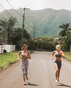Two women running barefoot on a road towards the camera with mountains in the background