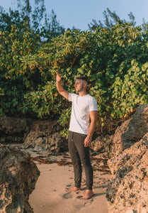 A man standing barefoot on the beach with greenery behind him wearing a white shirt and black joggers