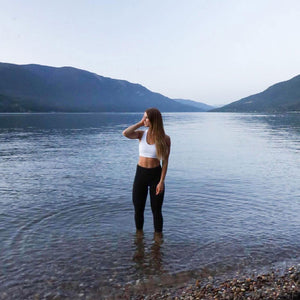 A woman standing in calm water close to a rocky beach with mountains behind her