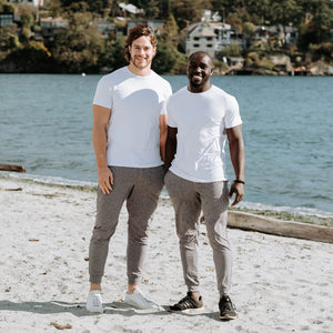two men standing on the beach wearing white neptune shirts and heather poseidon joggers