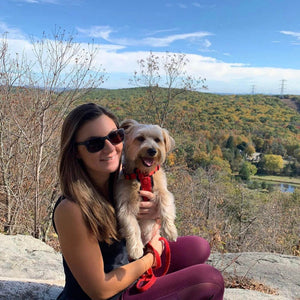 A woman sitting on a rocky cliff holding a small dog and wearing sunglasses and mesh leggings