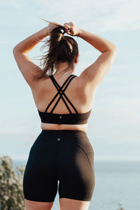 A woman wearing bike shorts and a sports bra looking out over the ocean and tying her hair back in a ponytail