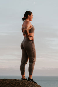 A woman wearing heather joggers and a sports bra standing on a rock overlooking the ocean